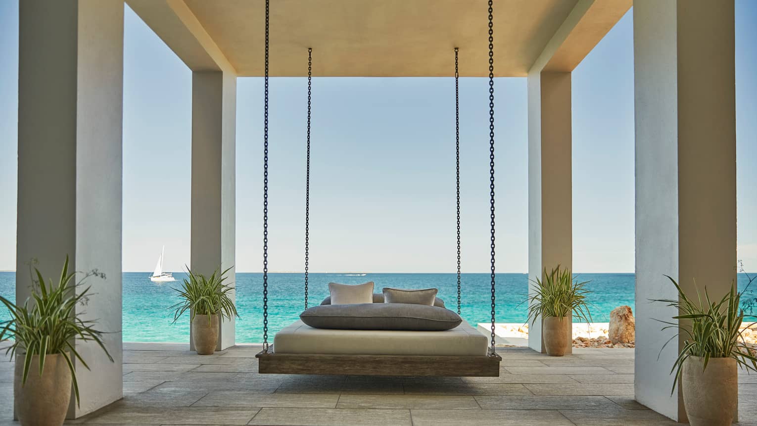 Patio bed hangs by chains from white ceiling in outdoor room with tropical potted plants, ocean view