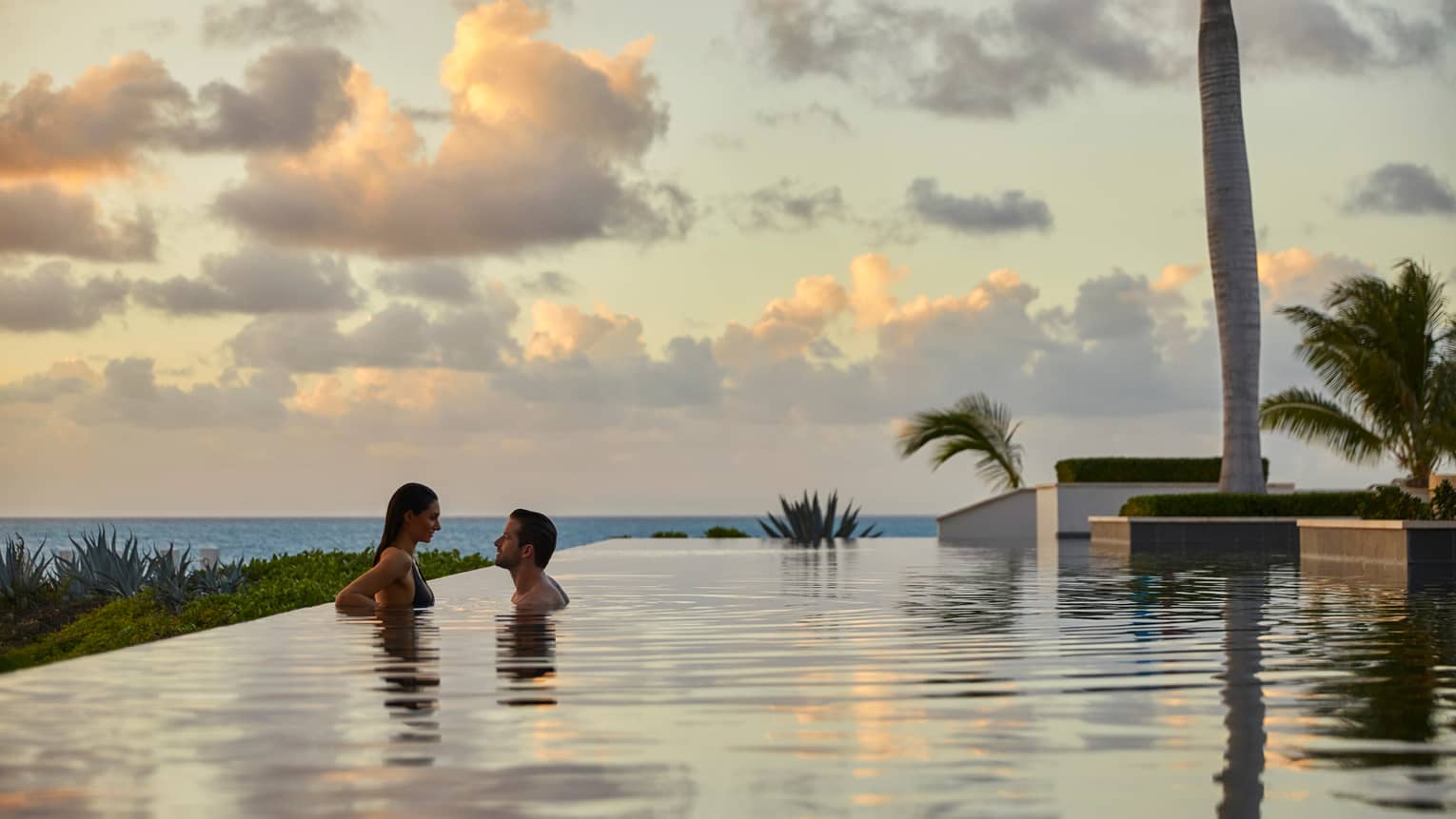 Couple wades in large infinity swimming pool under pink clouds, sunset 