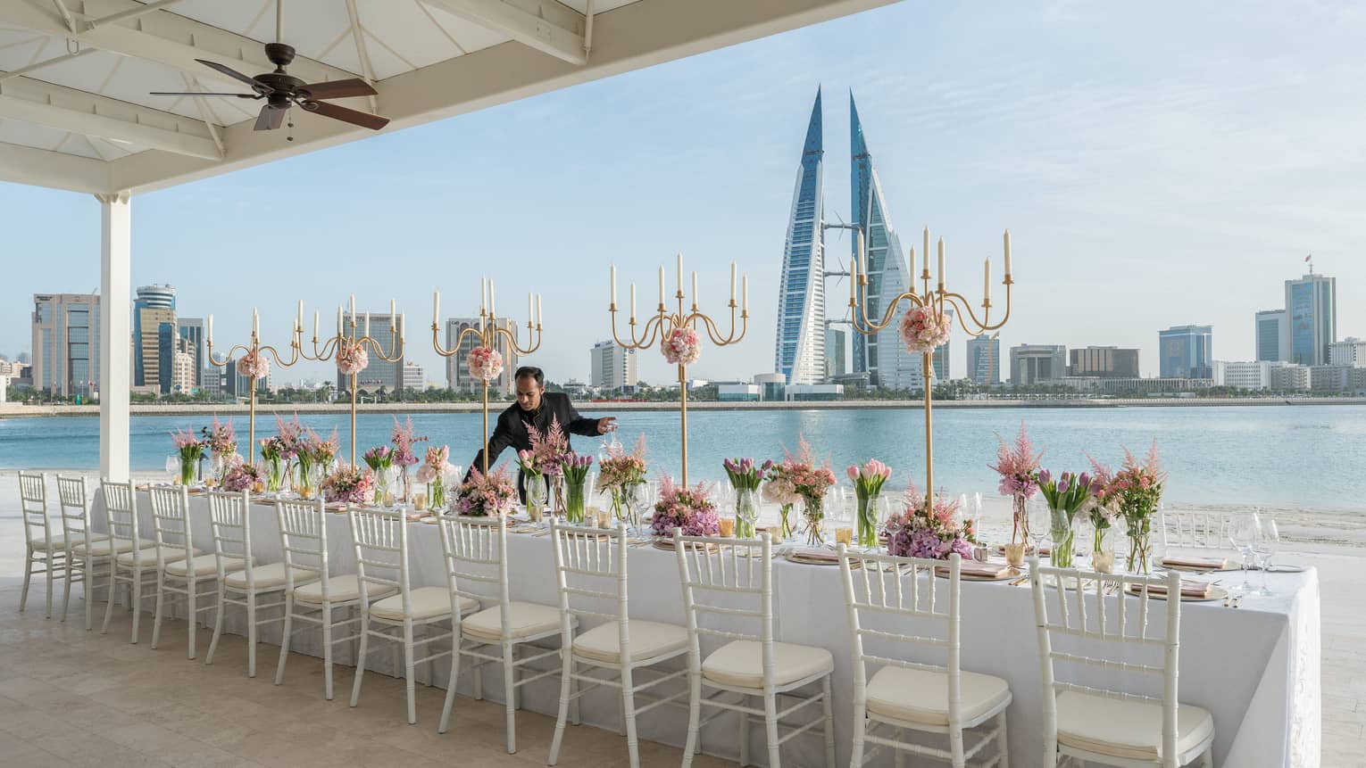 Hotel staff sets long wedding dining table by waterfront, city skyline in background