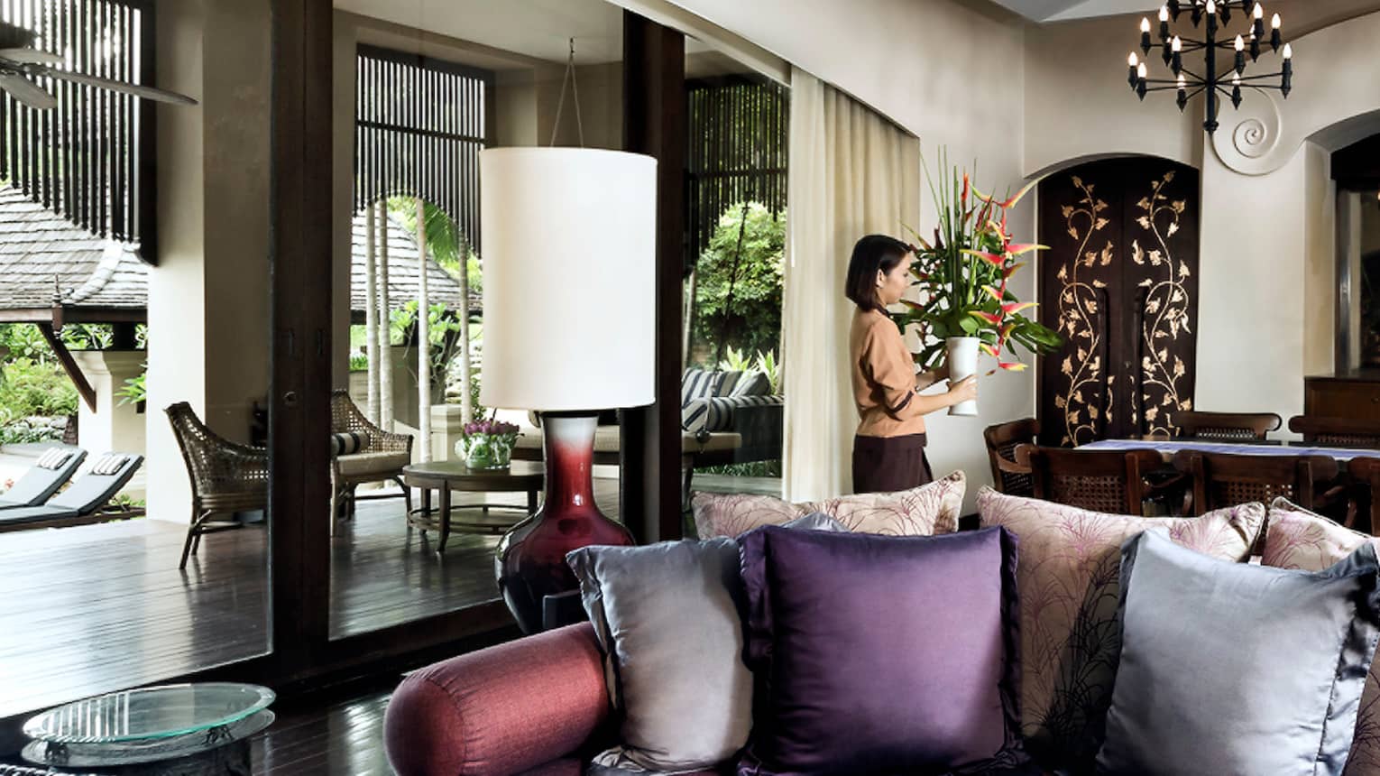 Hotel staff carries vase with tall tropical flowers past sofa, table in residence living room