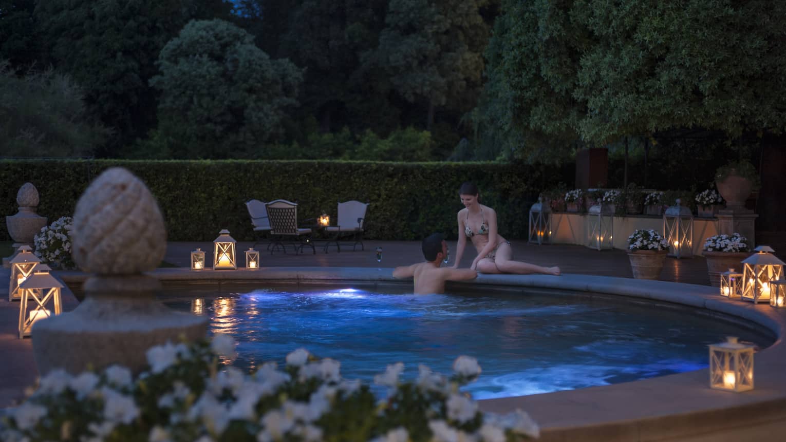 Woman sits on edge of outdoor whirlpool as man wades in water, glowing lanterns on deck