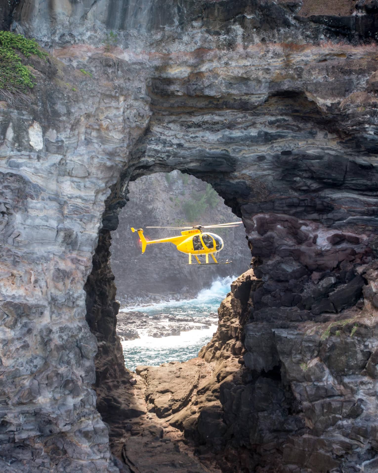 A sideview of a yellow holicopter, seen through an opening in a rock structure. 